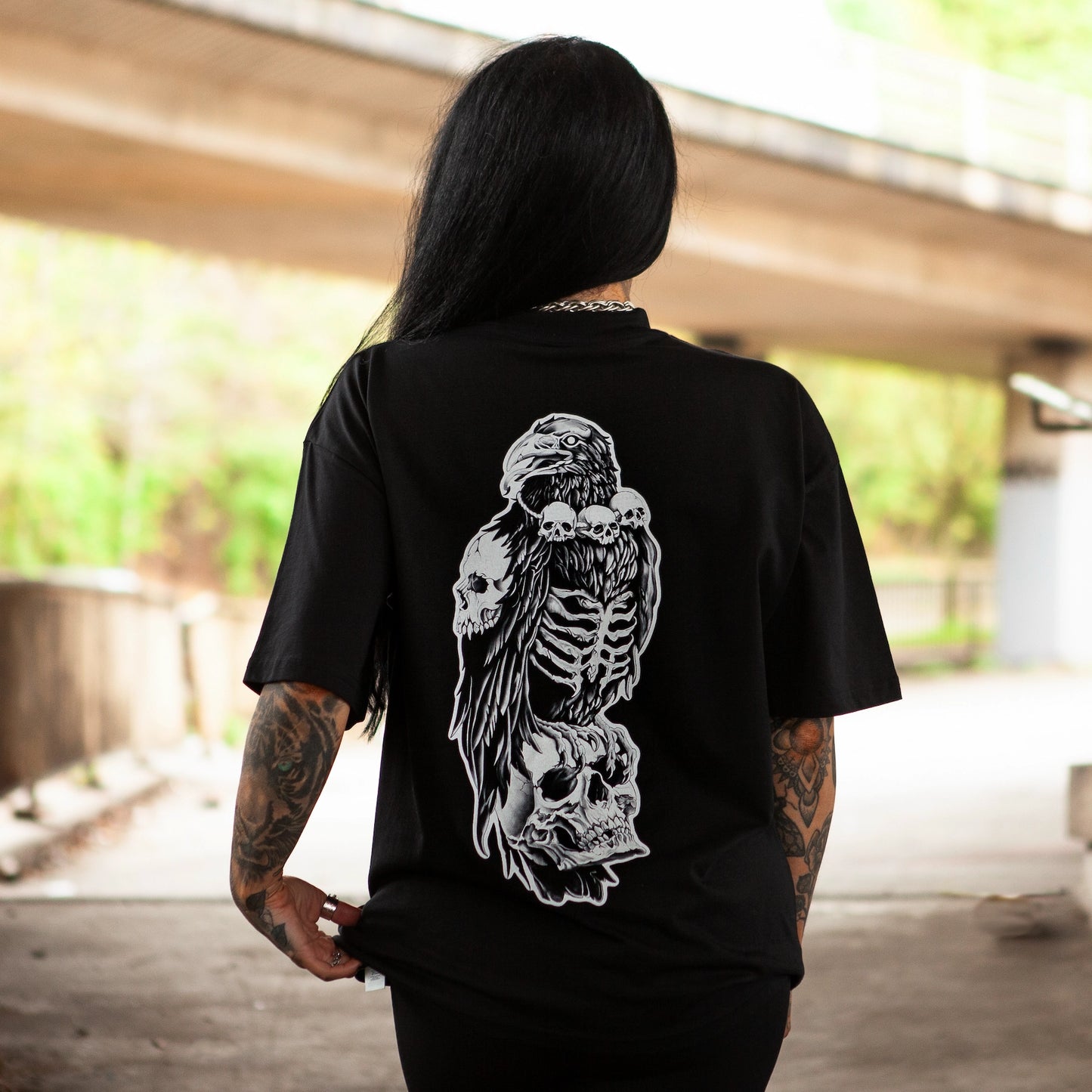 Back design of the Bone Collector t-shirt from Haunt Attire, showcasing a skeletal crow perched on stacked skulls in a white ink tattoo style. Photographed in urban underpass setting, Bristol.
