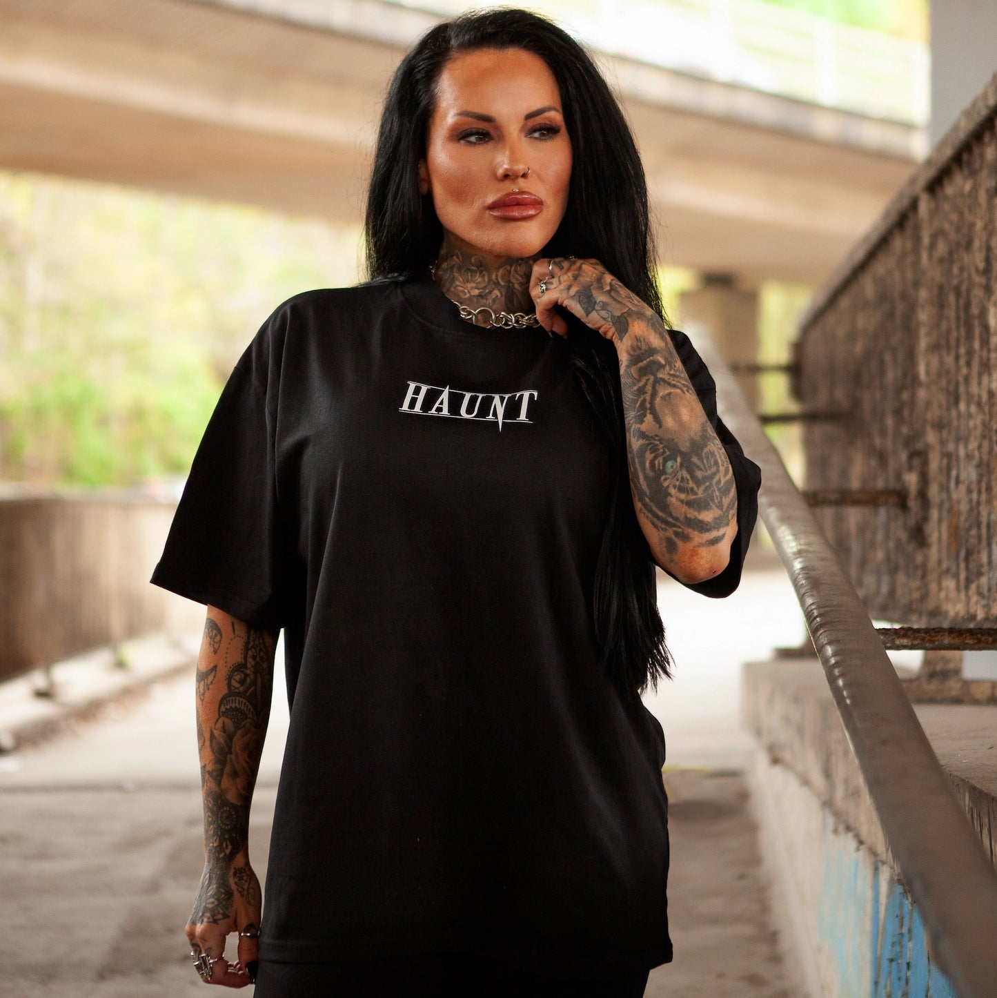 Front view of the Bone Collector premium oversized t-shirt by Haunt Attire, featuring white Haunt logo print on black tee. Shot under a concrete bridge in Bristol with tattooed model posing confidently.