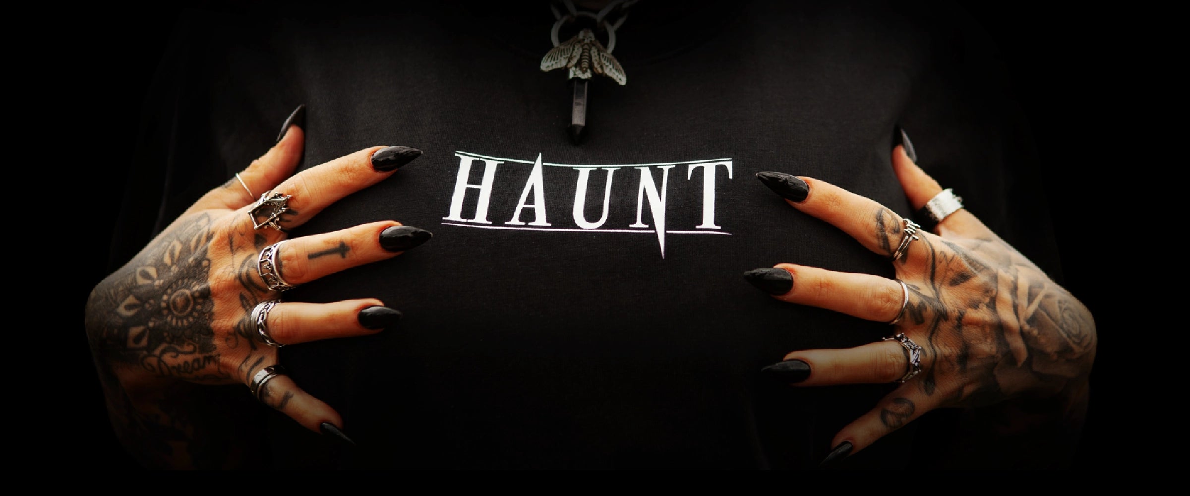 Close-up of the Haunt logo printed on a black t-shirt, framed by tattooed hands, silver jewellery and a moth pendant against a dark background.
