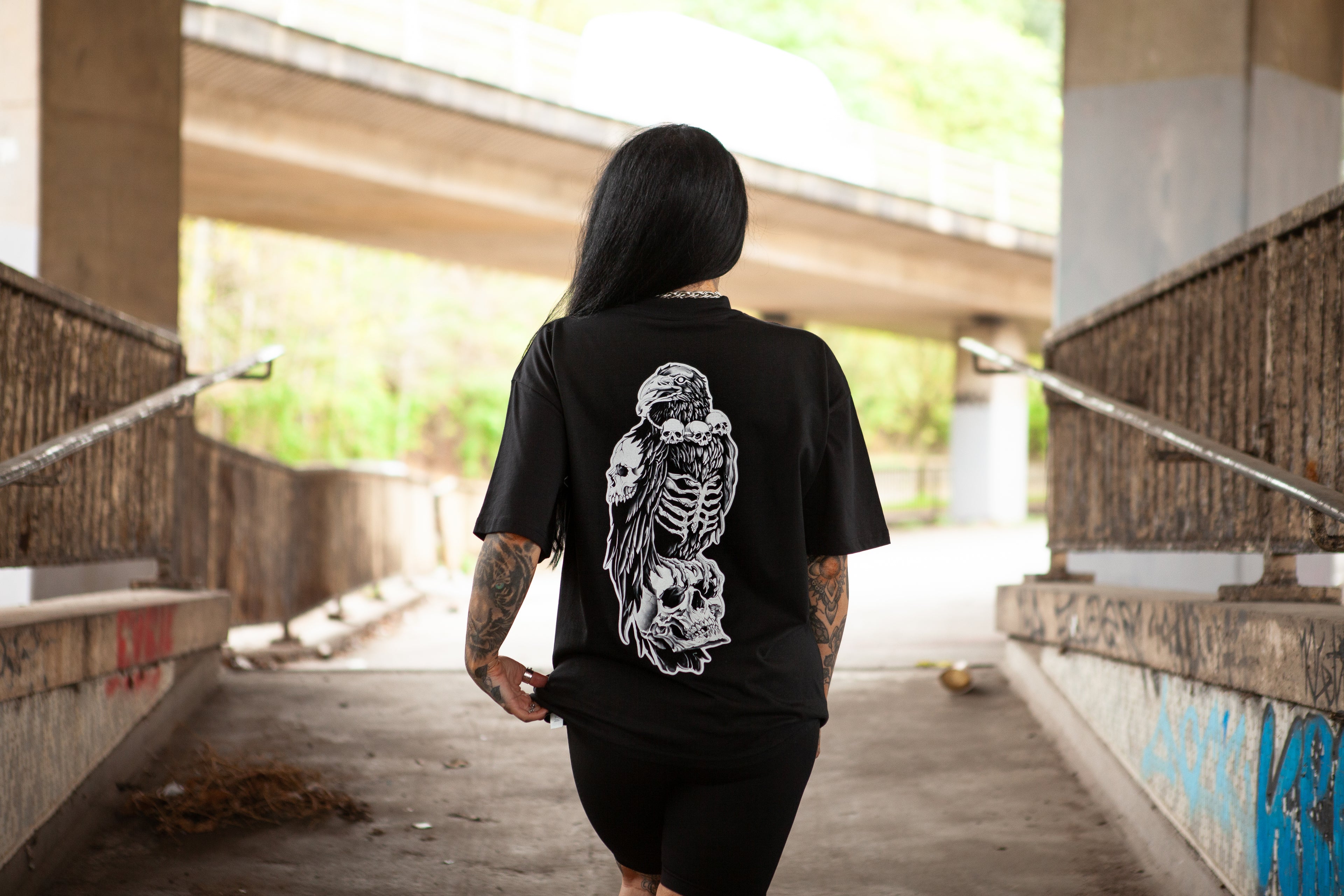 Back view of a person wearing a Haunt oversized t-shirt featuring the Bone Collector design with a raven and skull illustration, standing under a gritty urban underpass in Bristol.