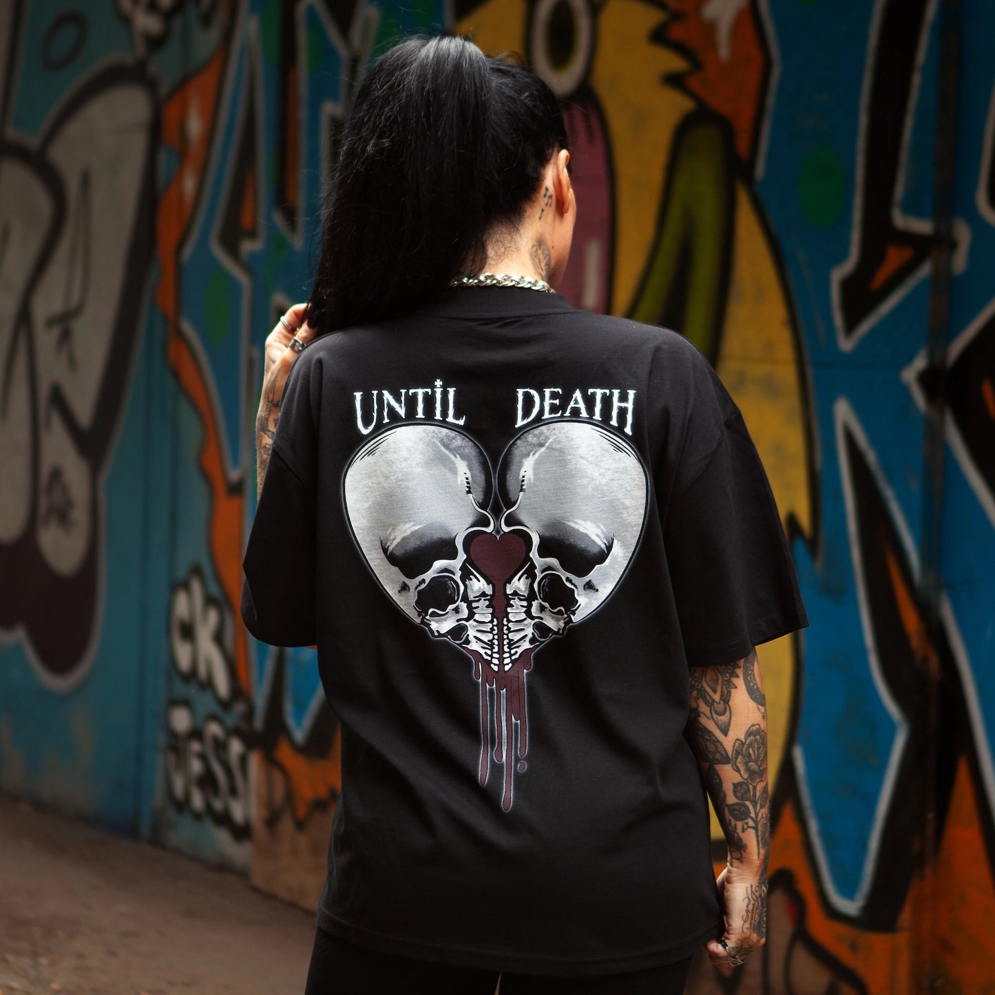 Back of Love Kills oversized tee, featuring twin skulls forming a heart with dark red drip, shot in a graffiti-covered alley.