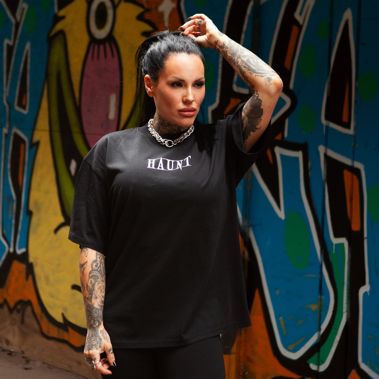 Model wearing black oversized Haunt tee with front HAUNT logo, posing in front of vibrant graffiti wall.