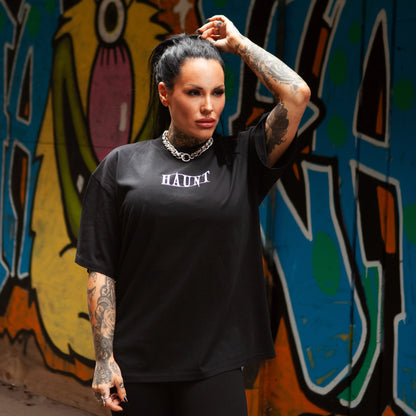 Model wearing black oversized Haunt tee with front HAUNT logo, posing in front of vibrant graffiti wall.