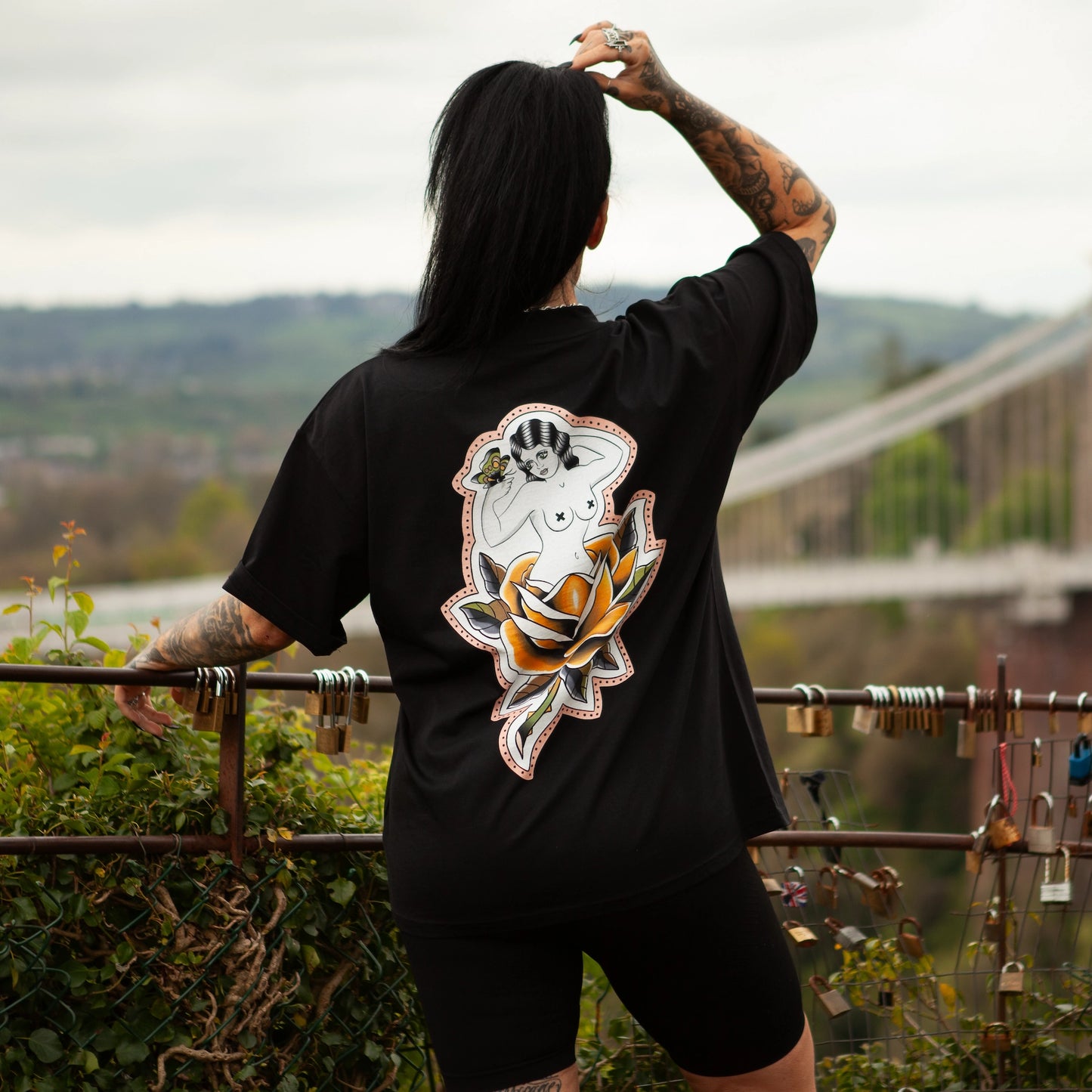 Model showing the back of the Softcore Bloom premium t-shirt, standing by the Clifton Suspension Bridge in Bristol with arm raised. Tattoo-inspired floral and a pin-up figure visible on black oversized shirt.