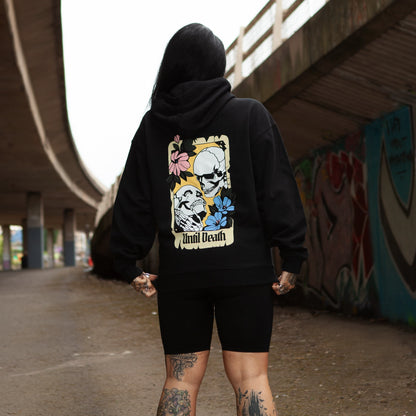 Model wearing the Until Death hoodie, back view under an urban underpass showing skull and flower artwork.